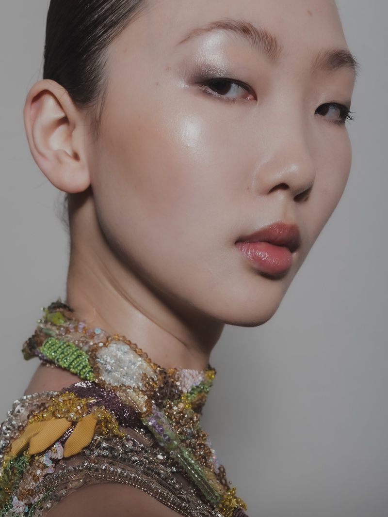 Close-up portrait of an Asian female model with flawless makeup and an ornate beaded collar, shot in a studio with neutral background.