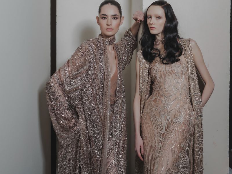 Two female fashion models pose indoors wearing elegant, embellished dresses and matching overcoats in a brightly lit photo studio setting.