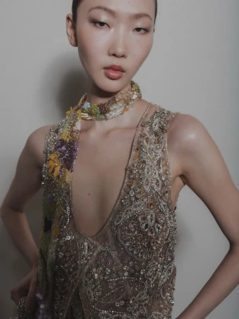 Model wearing an intricately embroidered sheer gown with beaded floral neckpiece against a neutral background