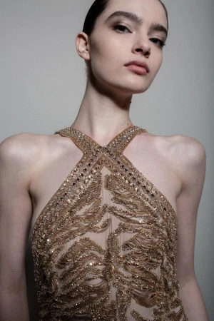 Model wearing a heavily embellished gold halter dress against a plain grey backdrop in a close-up fashion portrait