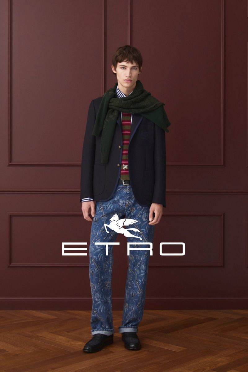A young adult male stands in a studio wearing a dark blazer, patterned jeans, striped shirt, and a green scarf, showcasing fashion.