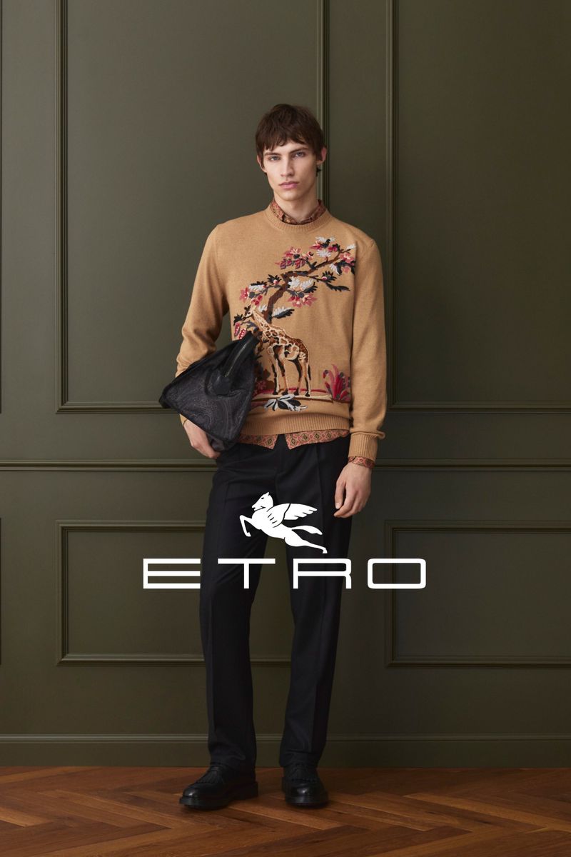 A young man poses indoors wearing a tan sweater with a giraffe design, black pants with an Etro logo, and black shoes.