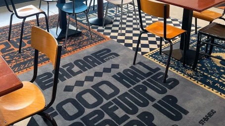 Interior view of a food hall featuring wooden dining chairs, tables, and patterned carpets with branded text under natural light.