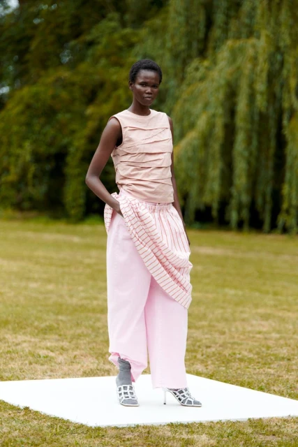An adult Black female fashion model poses in pink pants and a layered top on a white platform, outdoors with green trees in the background.