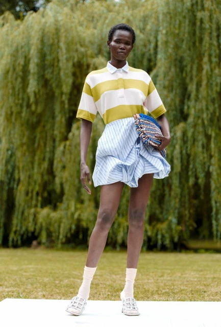 Full-length shot of a Black woman in a striped top and skirt, holding a clutch bag in an outdoor setting with green trees.
