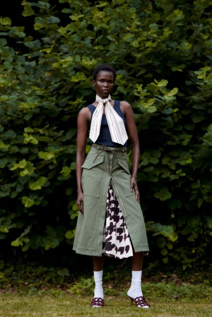 Woman in a sleeveless top and midi skirt with an open front panel stands in front of green foliage in a fashion shot.