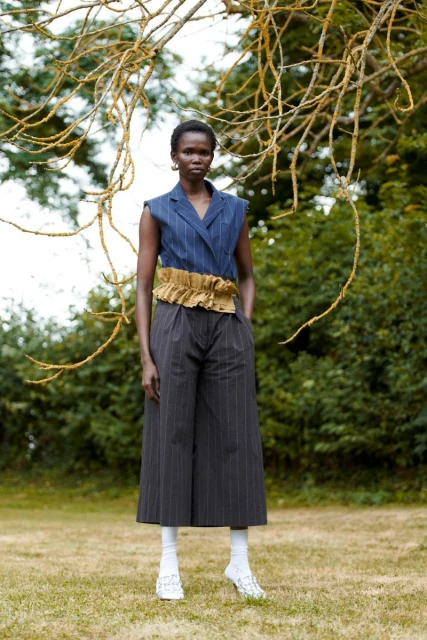 A slender Black woman poses in a sleeveless blue top, wide-legged pinstripe pants, and white socks with sheer shoes in a grassy outdoor setting.