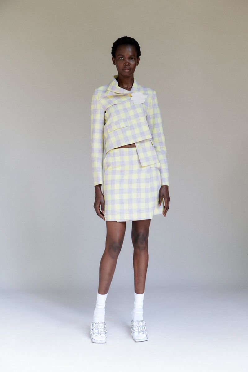 A full shot of a Black female model in a yellow and gray plaid jacket and skirt, white socks, and silver shoes, posing against a plain background.