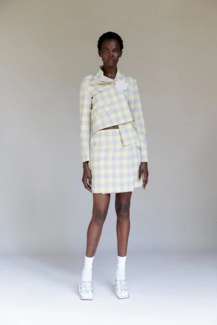 A full shot of a Black female model in a yellow and gray plaid jacket and skirt, white socks, and silver shoes, posing against a plain background.