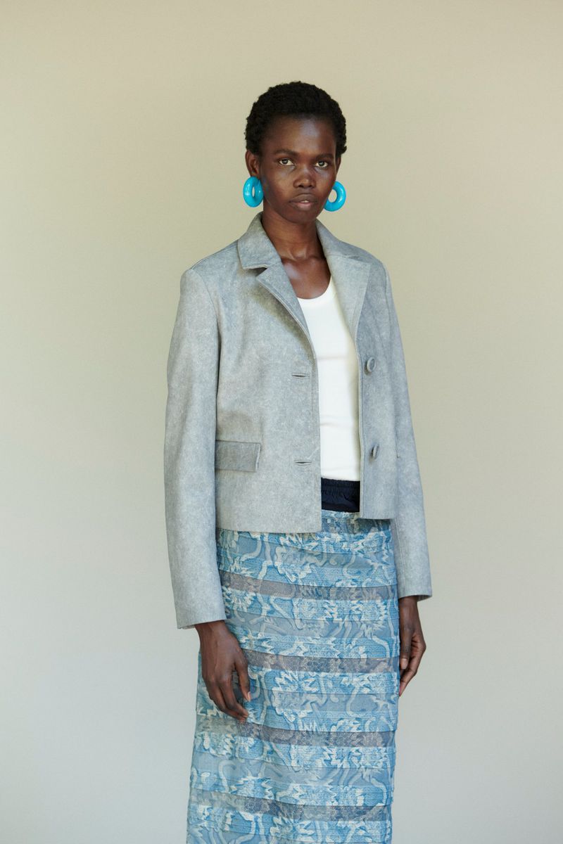 A slender Black woman in a grey blazer and blue skirt stands indoors with neutral expression, wearing large, round blue earrings.
