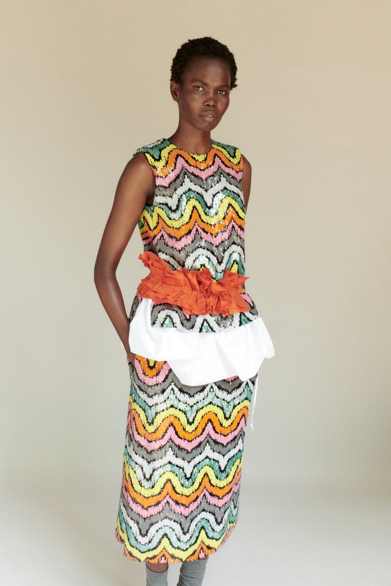 A dark-skinned woman poses in a sleeveless top and midi skirt with a wavy, zigzag pattern, orange fabric detail, and gray socks.