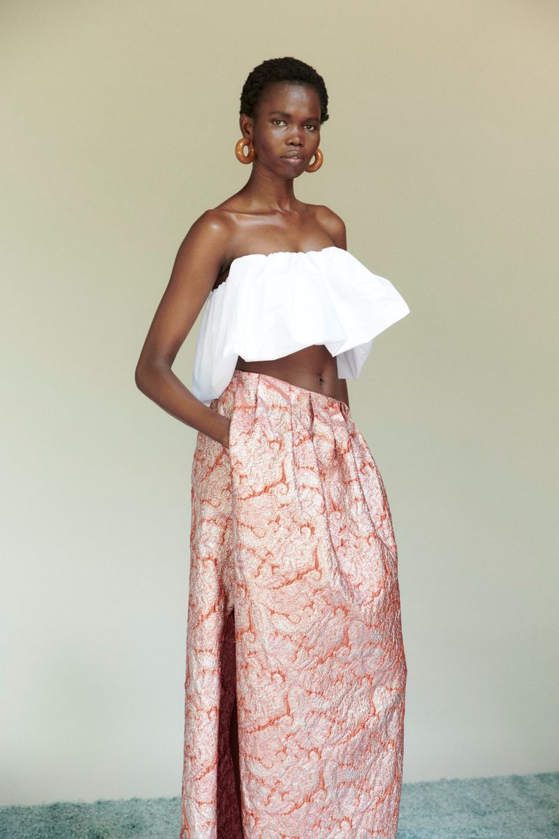 Young Black woman in a white tube top and patterned maxi skirt poses in a neutral studio setting, fashion editorial shot.