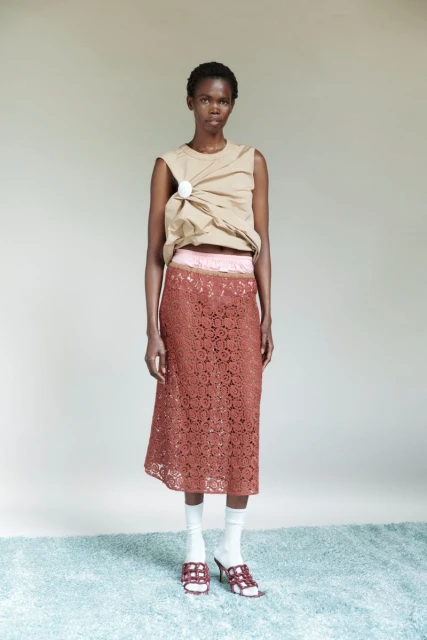 Full-length studio shot of a Black woman posing in a sleeveless beige top, long floral lace skirt, white socks and heeled sandals.