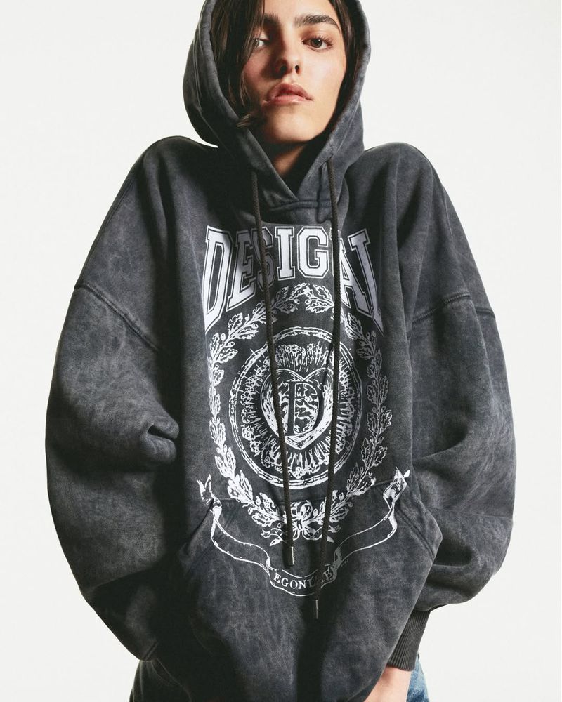 Woman in a gray hooded sweatshirt with a graphic design. She is in a studio setting.