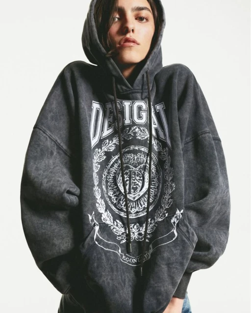 Woman in a gray hooded sweatshirt with a graphic design. She is in a studio setting.