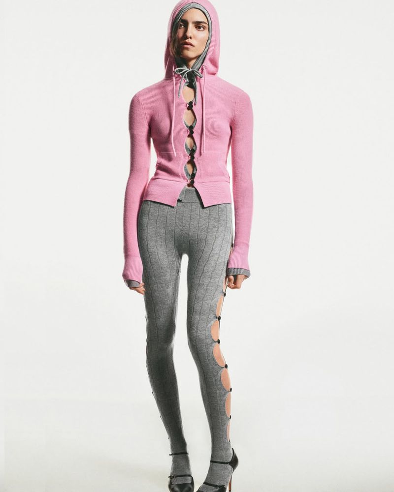 Woman in pink hoodie and grey patterned leggings posing in a full shot on a white background in a fashion editorial studio photograph.
