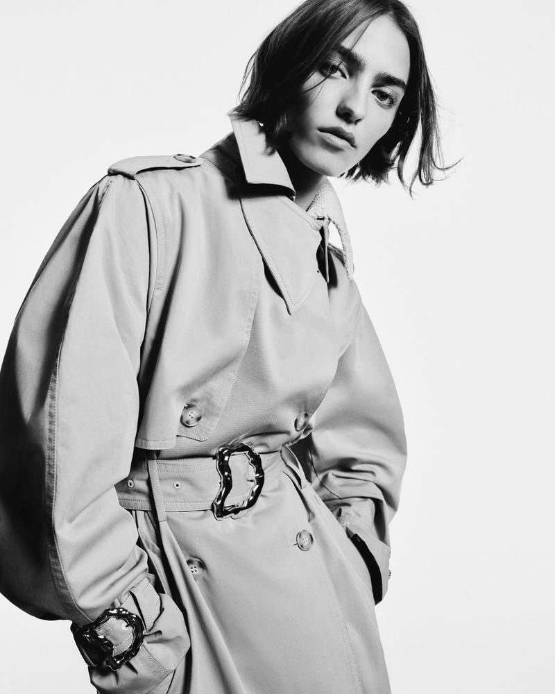 Monochrome portrait of a woman in a beige trench coat with oversized belt buckle, looking at the camera against a light background.