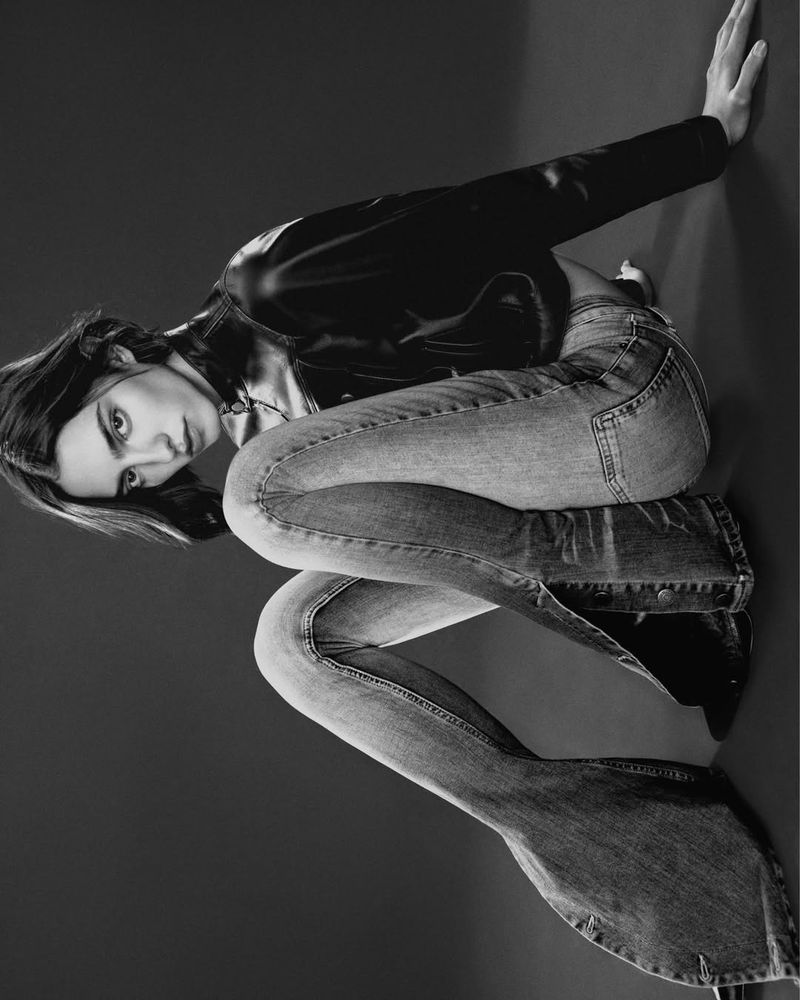 Monochrome portrait of a young adult woman in a black leather jacket and jeans, posing in a photo studio with dynamic lighting.