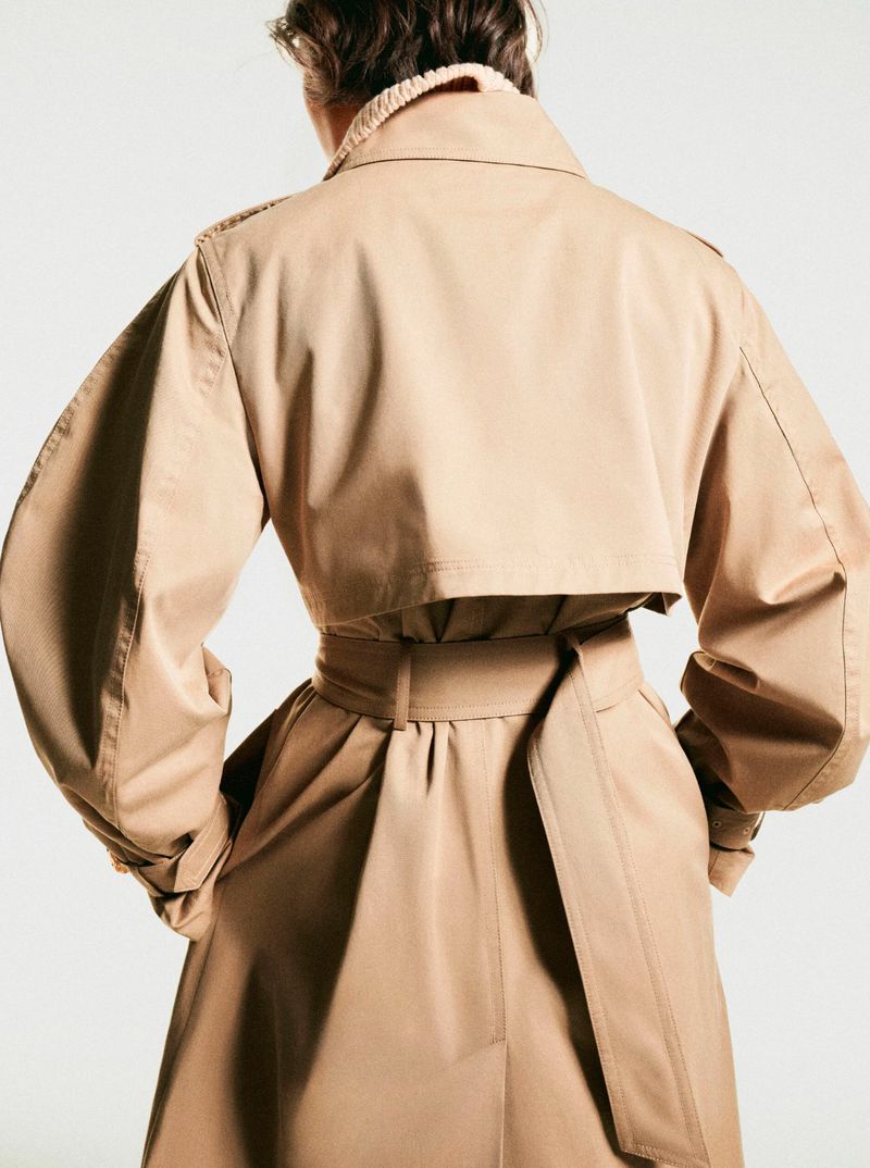 Model from behind wearing a tan trench coat with a belted waist and voluminous sleeves against a plain white background