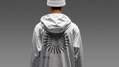 Person wearing a white hooded jacket with a black graphic on the back and a white beanie against a grey background.