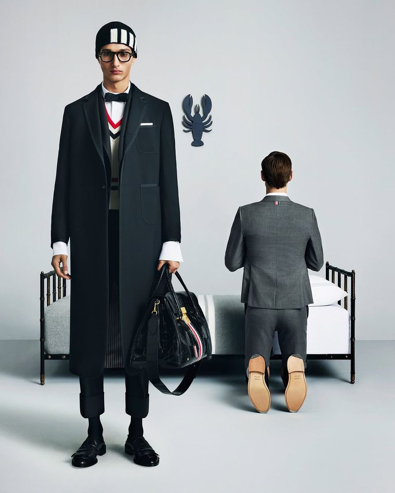 Model in dark coat and beanie holding a black bag stands beside a kneeling model in a gray suit before a bed.