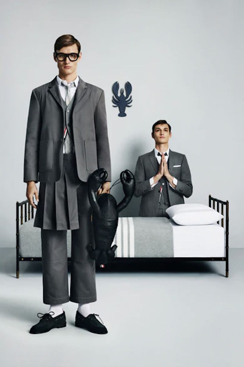 Two male models in gray suits pose in a photo studio; one model holds a black lobster prop, and a bed and lobster art hang behind them.