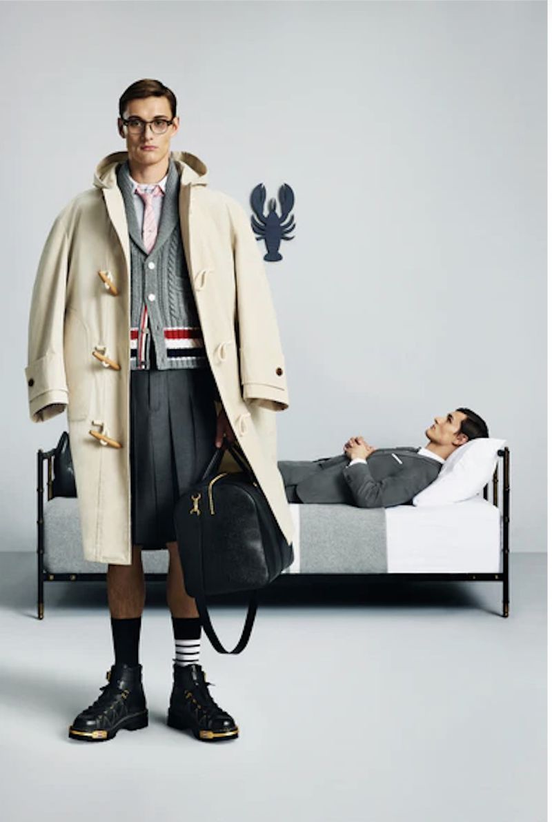 Two adult men in a studio setting; one stands in an oversized coat and pleated shorts, while the other relaxes on a bed. 