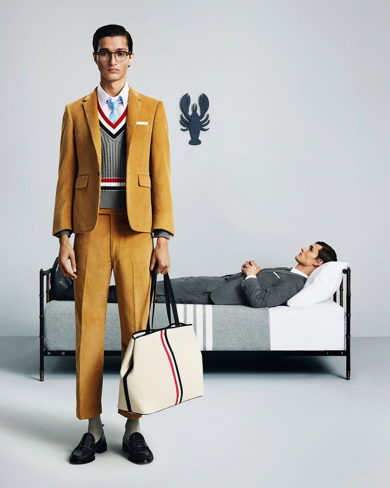 A male fashion model stands in a gold suit with a tote bag, another model reclines on a bed in a grey suit in a studio.