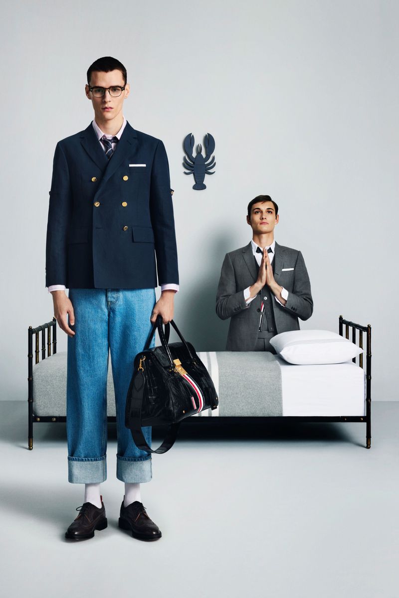 Two men in suits in a surreal, fashion-forward studio setting. One is standing holding a duffel bag, the other is near a bed.