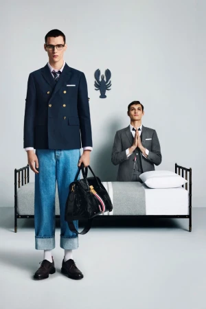 Two men in suits in a surreal, fashion-forward studio setting. One is standing holding a duffel bag, the other is near a bed.
