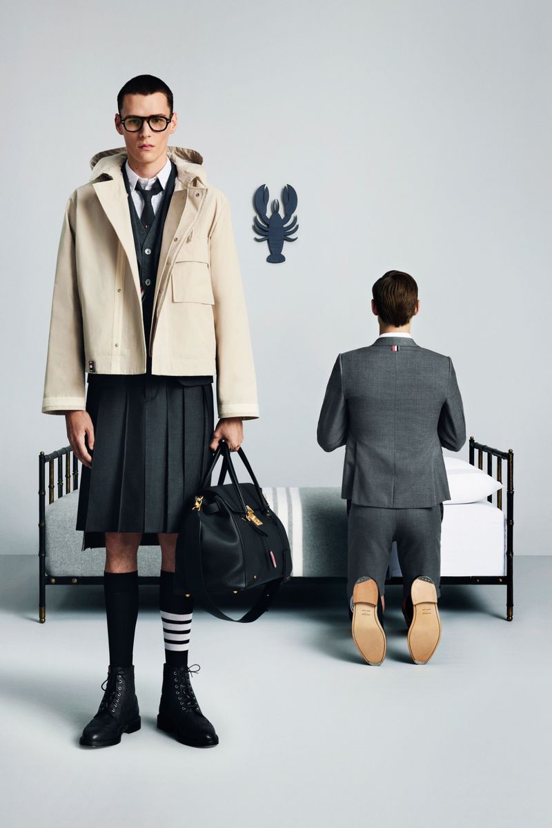 Two adult men in a studio setting. One man stands wearing a kilt and jacket, and holding a bag. The other man kneels on a bed.