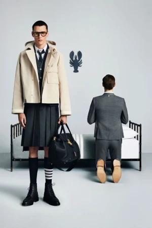 Two adult men in a studio setting. One man stands wearing a kilt and jacket, and holding a bag. The other man kneels on a bed.