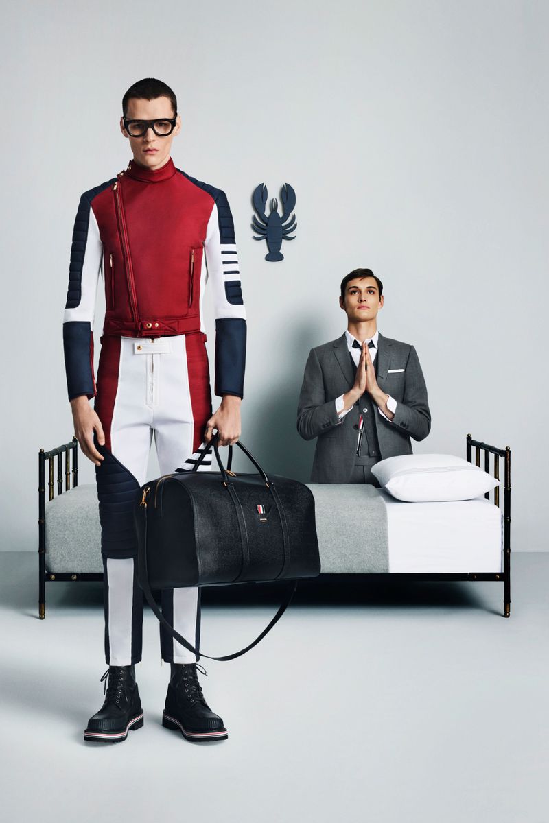 Editorial fashion shot of two adult men in a photo studio, one standing with a duffel bag, the other kneeling on a bed.