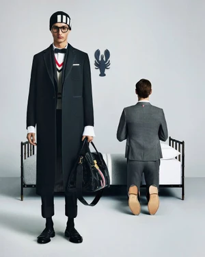 Model in dark coat and beanie holding a black bag stands beside a kneeling model in a gray suit before a bed.