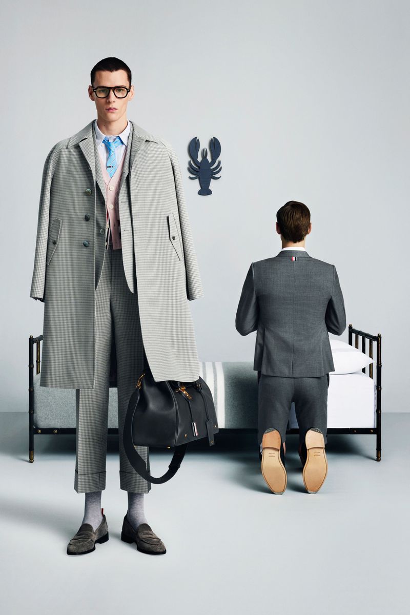 Two male models in grey Thom Browne tailored suits, one standing holding a black leather bag and one kneeling near a bed