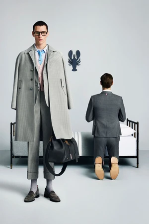 Two male models in grey Thom Browne tailored suits, one standing holding a black leather bag and one kneeling near a bed
