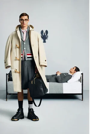 Model wearing cream duffel coat, grey shorts, and carrying a black duffel bag with another man lying on a bed in the background.