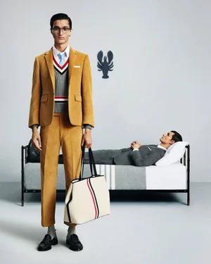 Model in a mustard yellow suit holding a tote bag with a second model lying on a bed in the background against a light grey wall