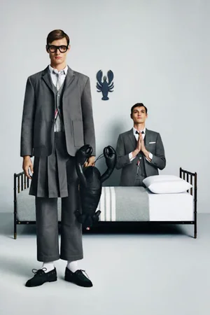 Two men in grey Thom Browne suits, one standing holding a leather lobster prop and another seated behind a bed.