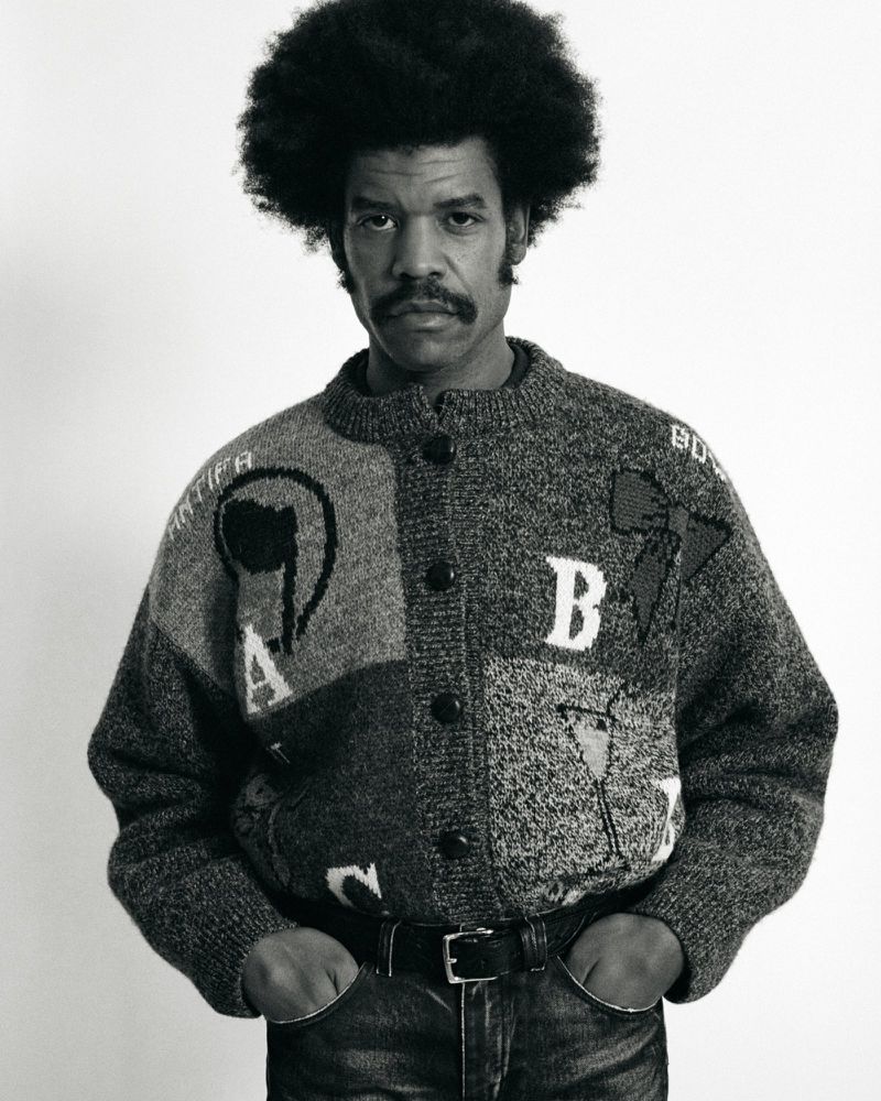 Monochrome studio portrait of a man with an afro, wearing a patterned cardigan, jeans and belt, hands in pockets, against a white backdrop.