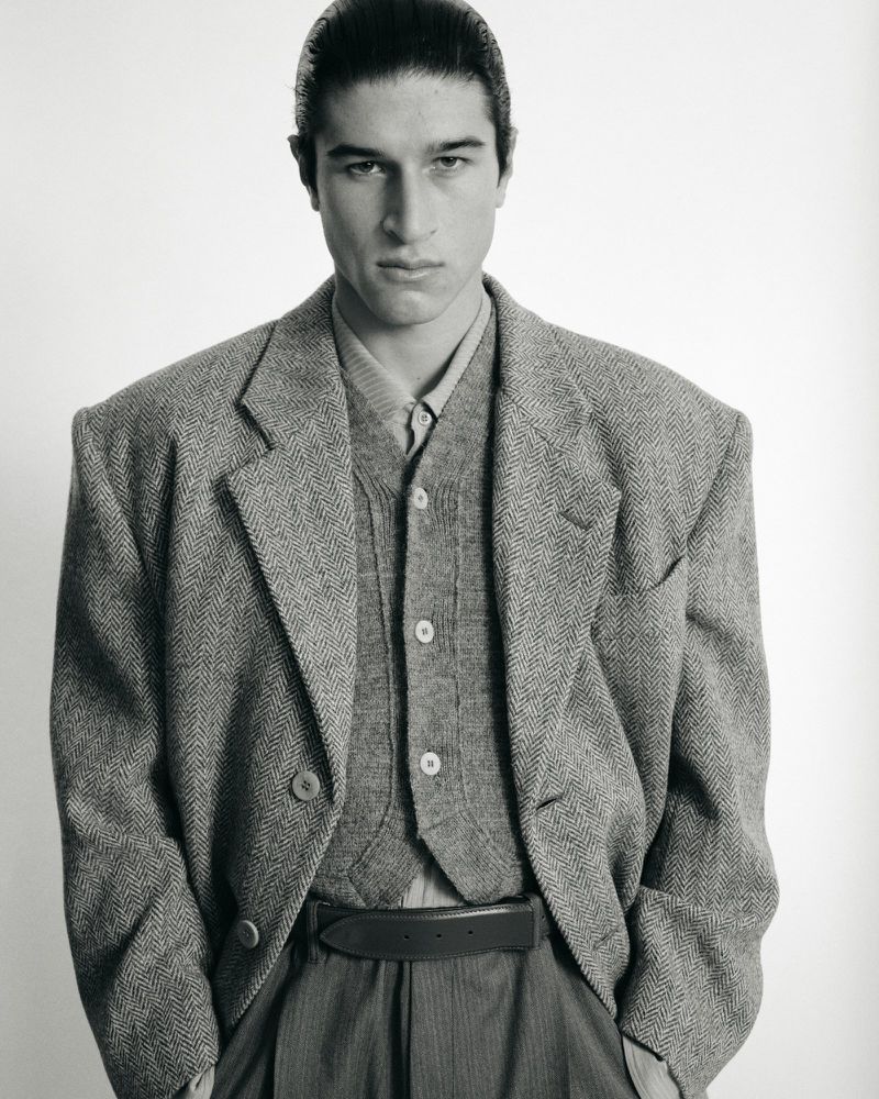 Monochrome portrait of a man in a grey tweed blazer and vest looking at the camera against a white background, hands in pockets.