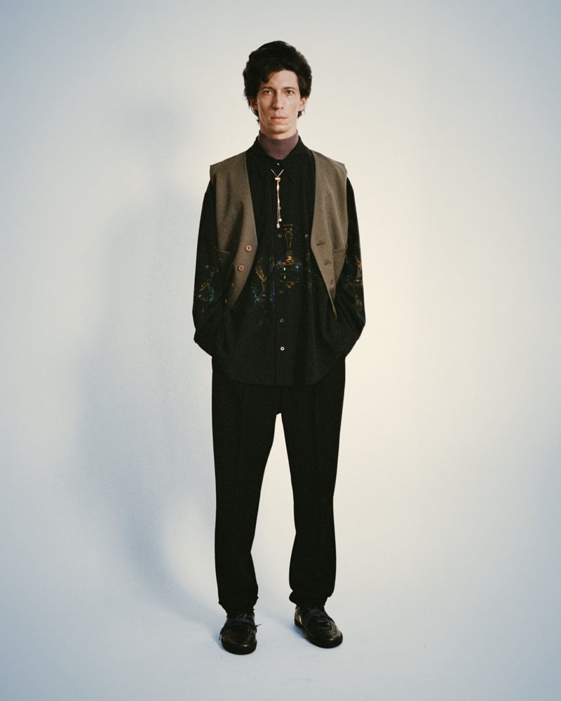 Portrait of a slender adult man with dark hair in a patterned shirt, vest, and black pants against a bright, neutral backdrop.