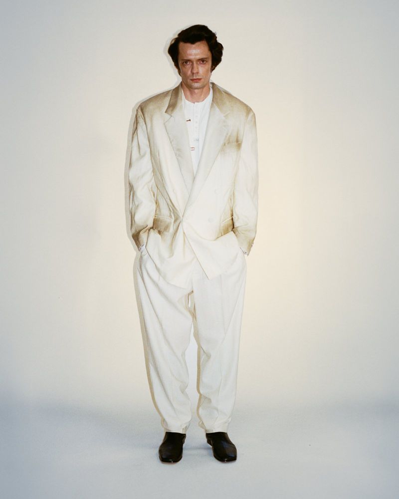 A full portrait shows a male model in a light-colored suit, standing against a bright background in a fashion or editorial setting.