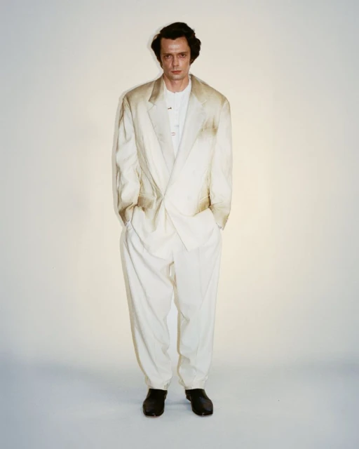 A full portrait shows a male model in a light-colored suit, standing against a bright background in a fashion or editorial setting.