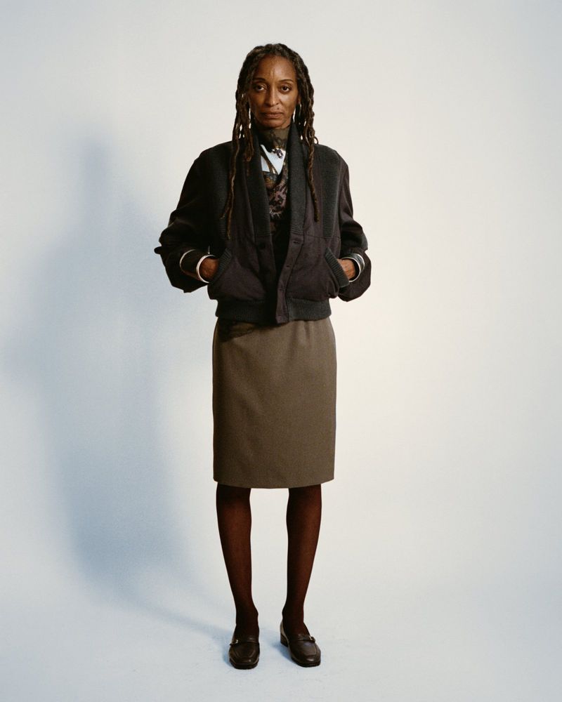 Full-length portrait of a Black woman with dreadlocks in a brown skirt and black jacket, standing in front of a white background.