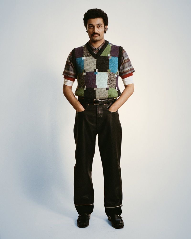 Full-length portrait of an adult male wearing a patterned sweater vest, dark trousers, and shoes against a plain white backdrop.