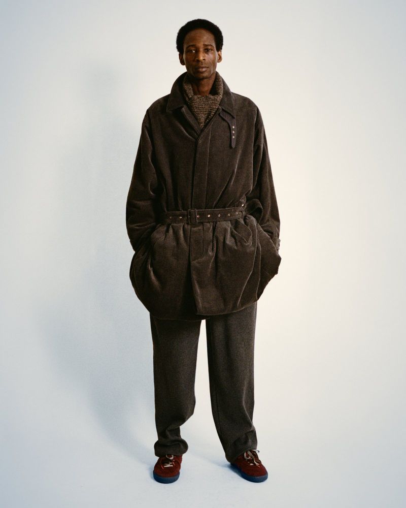 A Black male model poses in a brown corduroy jacket and trousers, with red and brown shoes, in a full shot against a plain white background.