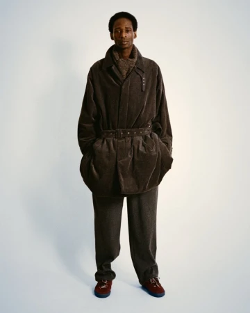 A Black male model poses in a brown corduroy jacket and trousers, with red and brown shoes, in a full shot against a plain white background.