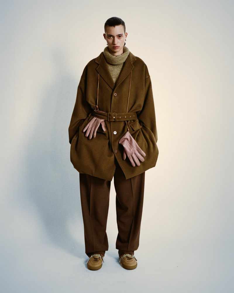 Fashion portrait of a young adult wearing a brown jacket with a belt, trousers, tan sneakers, and beige gloves in a studio setting.