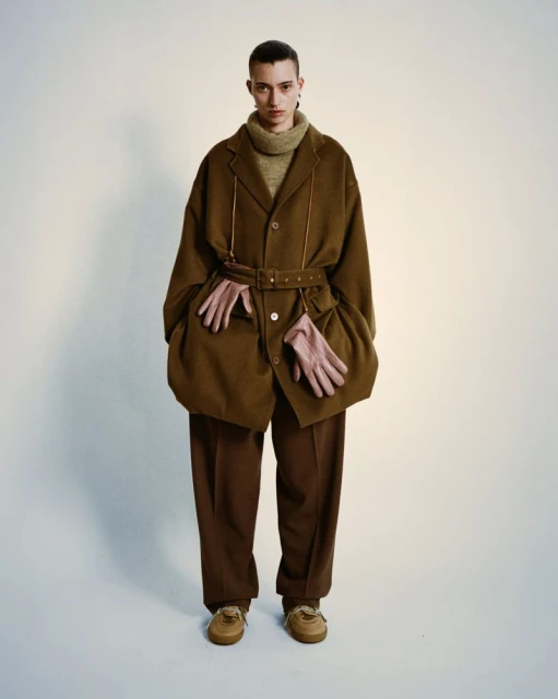 Fashion portrait of a young adult wearing a brown jacket with a belt, trousers, tan sneakers, and beige gloves in a studio setting.